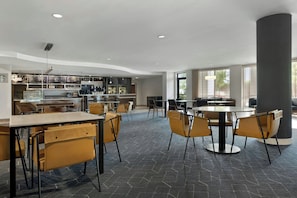 Un comedor moderno con mesas y sillas, una cocina al fondo y ventanas amplias.