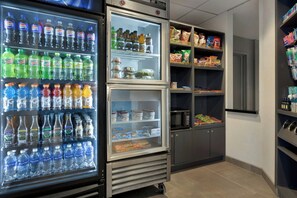Una máquina expendedora con diversas bebidas y aperitivos, un microondas y un refrigerador con varios estantes.