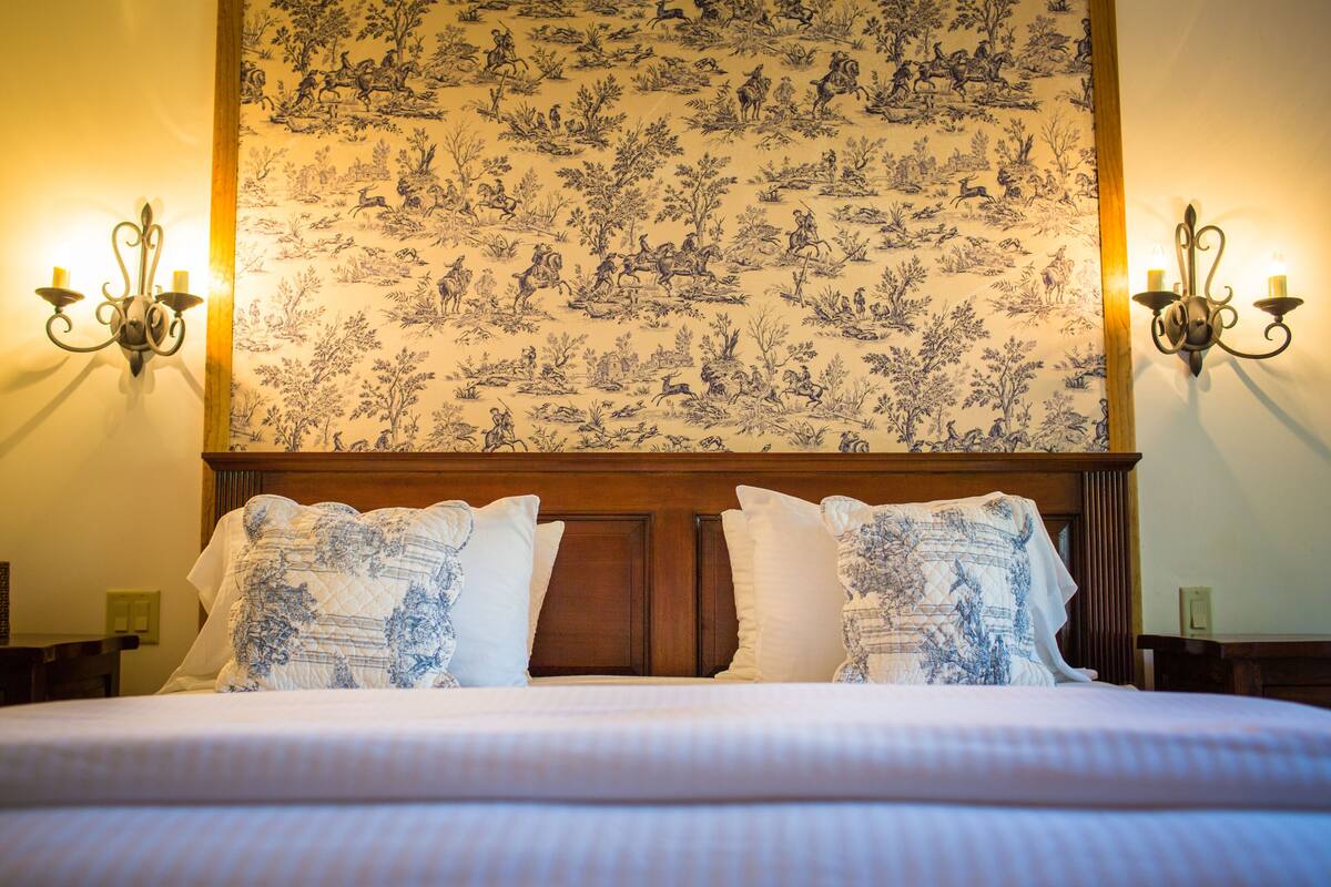 A bed with a floral-patterned headboard, two wall sconces, and a wall with a detailed tapestry.