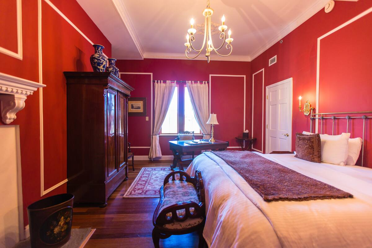 A bedroom with a large bed, a wooden dresser, a chandelier, and a window with curtains.