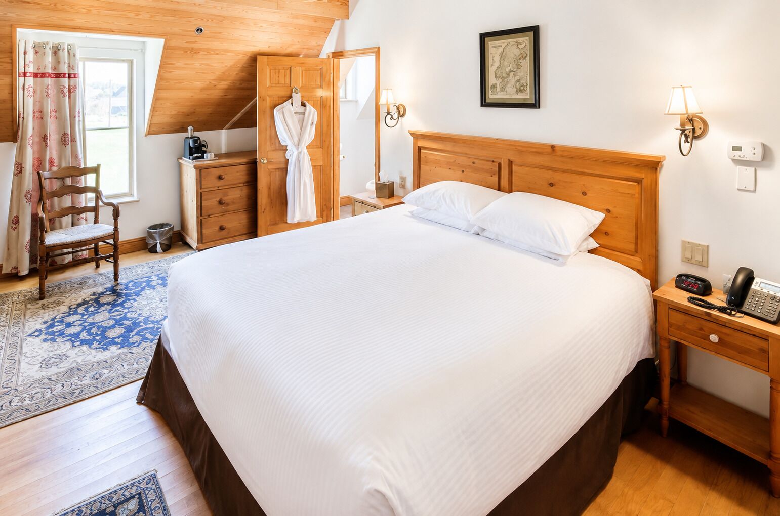 A hotel room with a large bed, wooden headboard, a bedside table with a phone, a nightstand, a wooden dresser, a robe, a chair, a window with curtains, and a wall-mounted lamp.