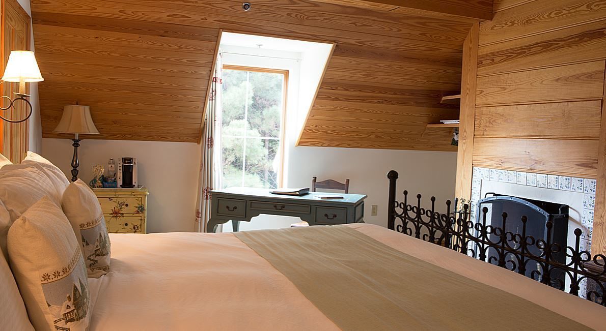 Un dormitorio con techo de madera, una cama con cubrecama verde, una mesita de noche con lámpara, un aparador y una ventana con vista a árboles.