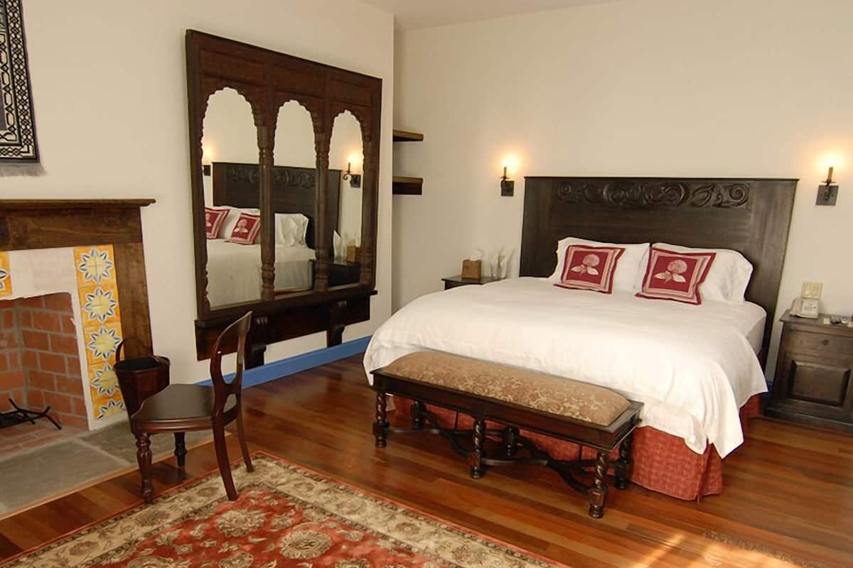 A bedroom with a large bed, a wooden bench, a mirror, and a fireplace.