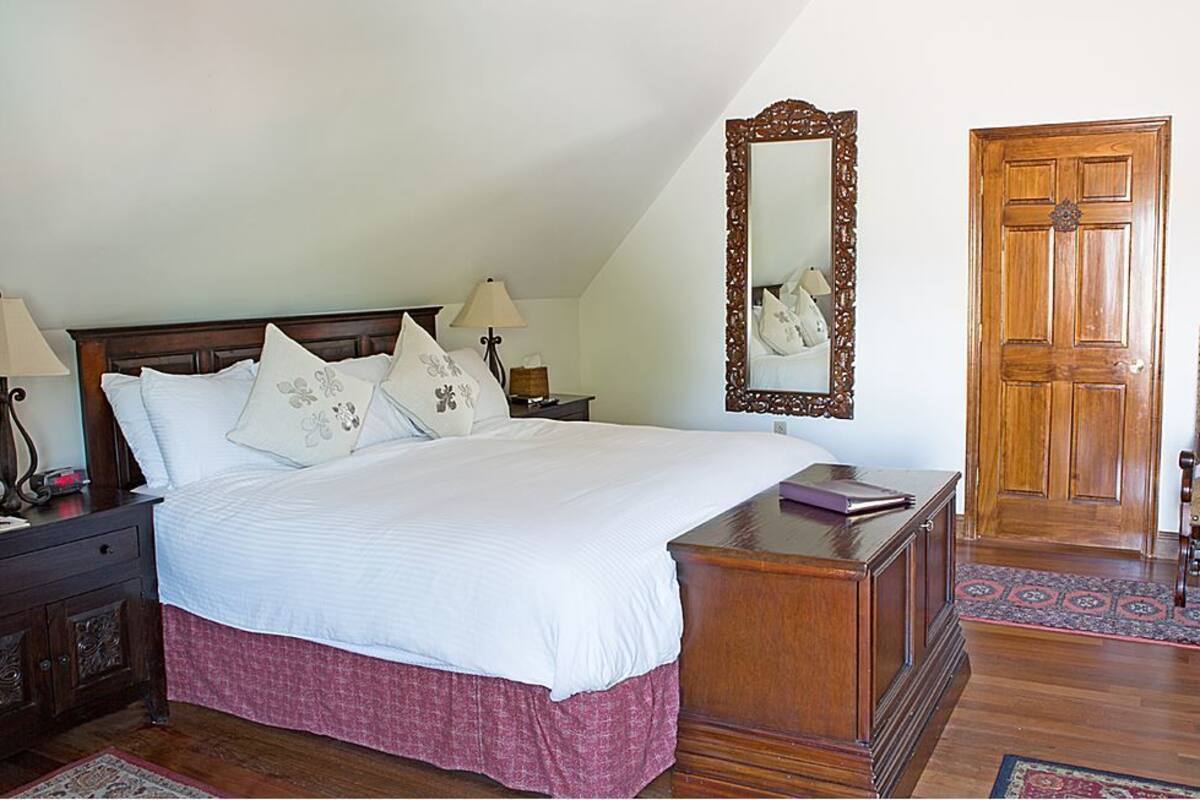 A bedroom with a large bed, wooden headboard, bedside tables with lamps, a mirror, a wooden door, and a chair with a cushion.