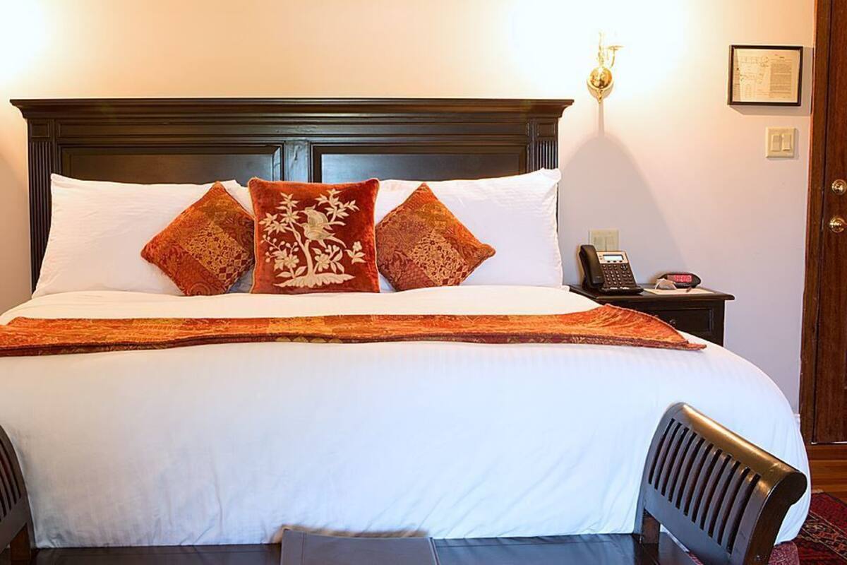 A neatly made bed with white linens and two decorative pillows, a bedside table with a telephone, and a wall-mounted light fixture.