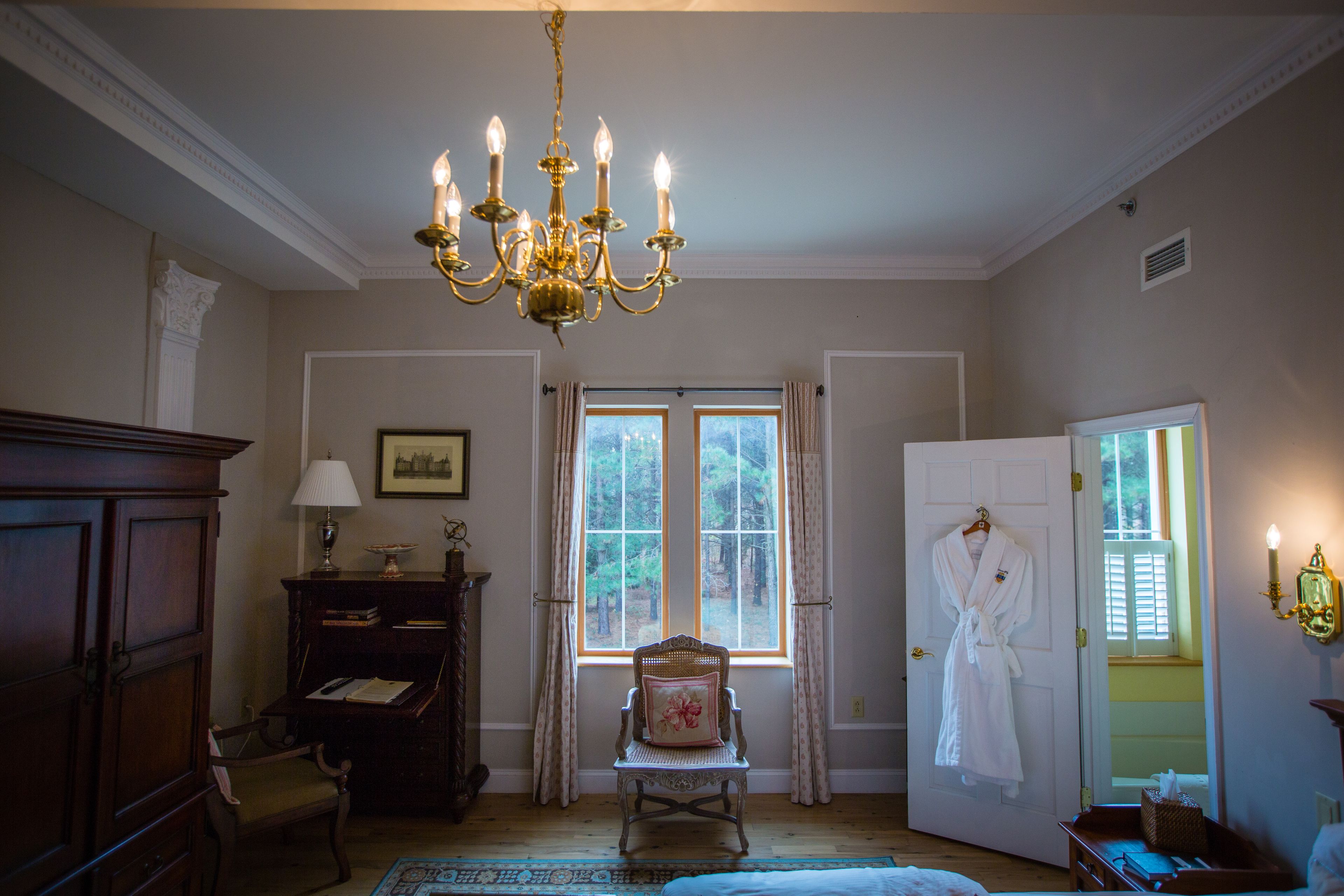 A room with a chandelier, a chair with a cushion, a wooden cabinet, a window with curtains, and a door with a robe hanging.