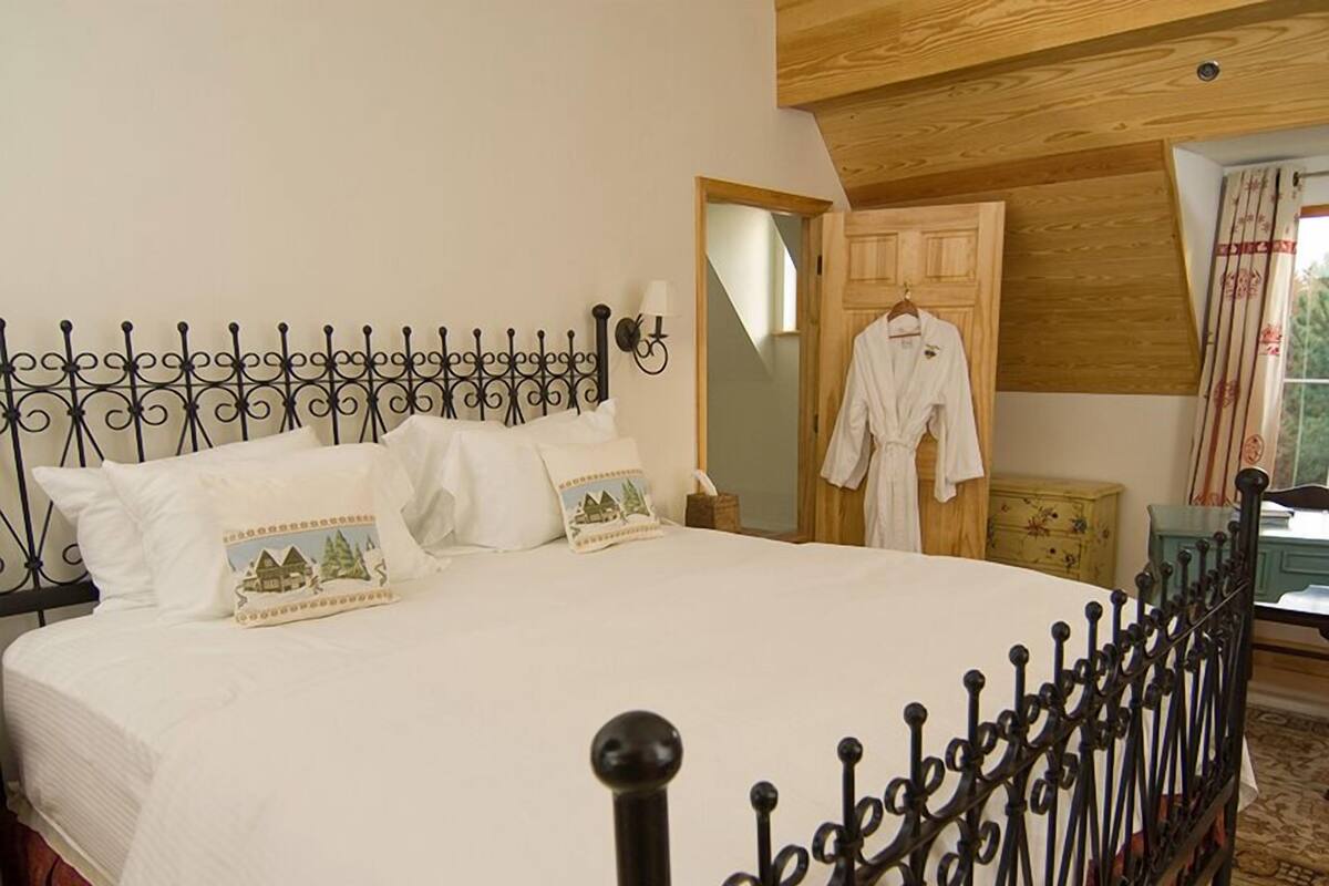 A neatly made bed with a metal headboard, white bedding, and decorative pillows. A wooden door and a robe hanging on a hook are visible in the background.