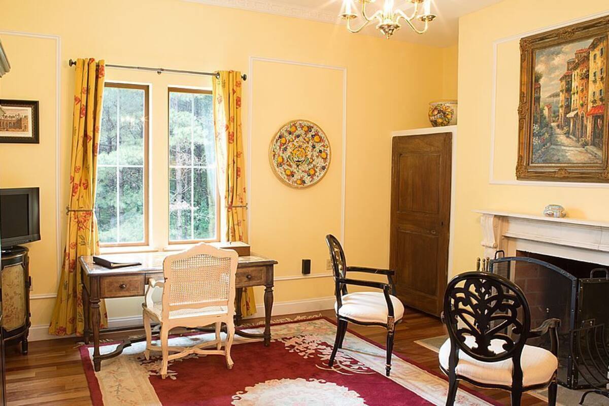 A room with a wooden desk, two chairs, a fireplace, and a large window with curtains.