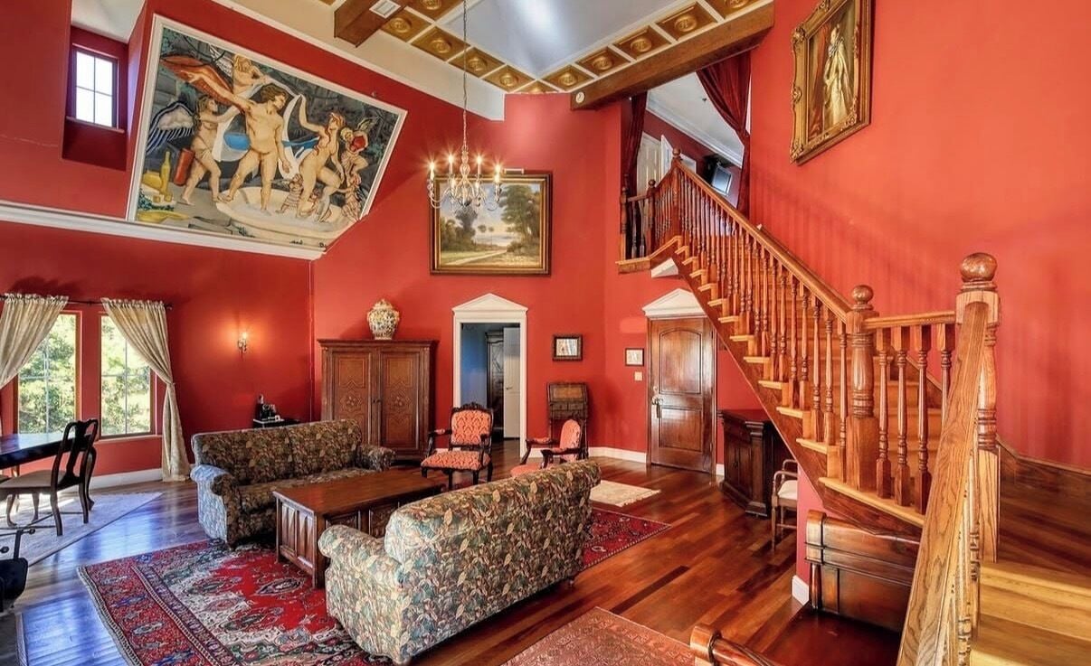 A spacious living room with a high ceiling, a wooden staircase, and a large painting.