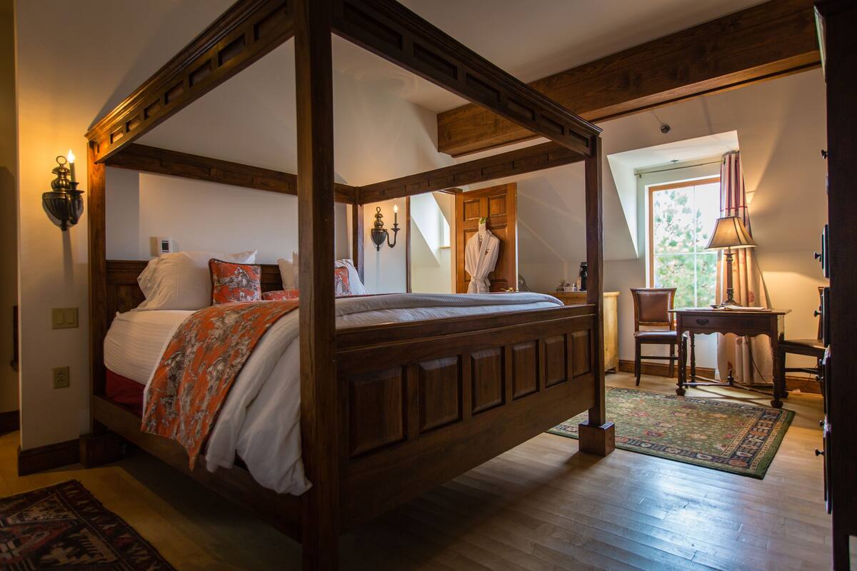 A four-poster bed with a wooden frame, a nightstand, a chair, and a desk with a lamp.