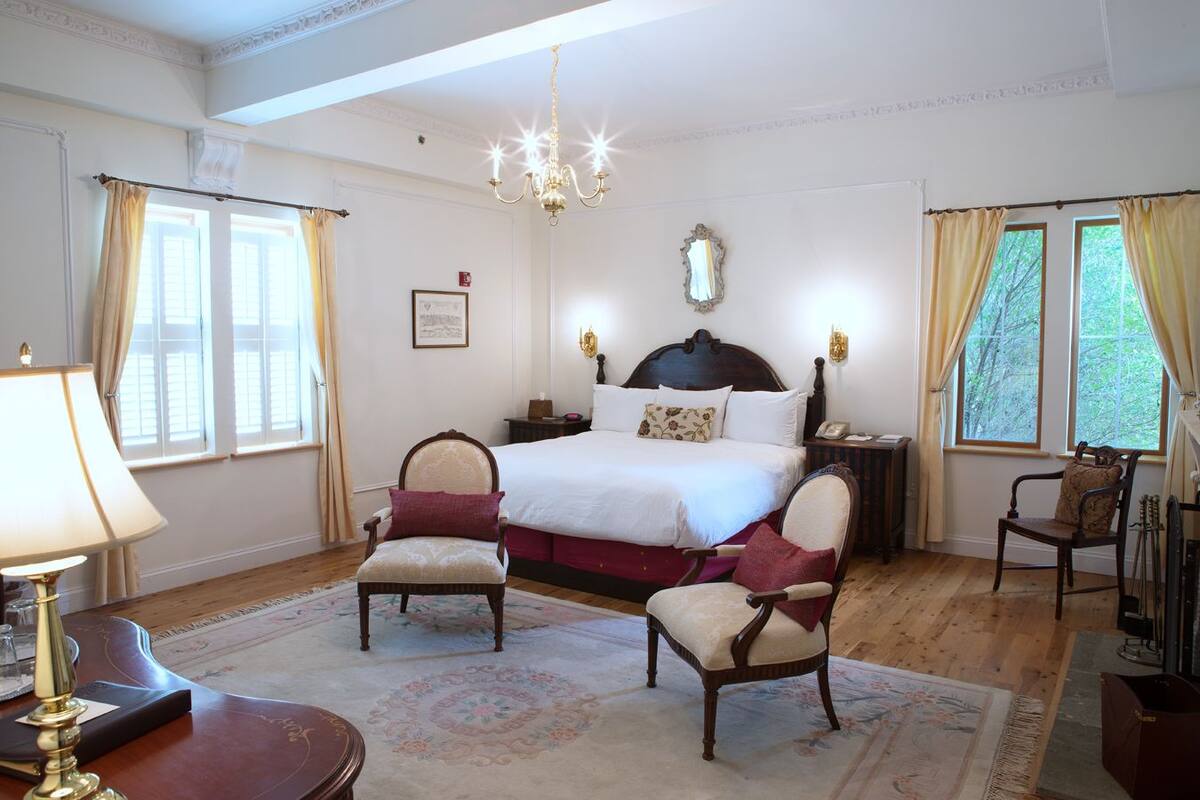 A spacious bedroom with a large bed, two chairs, a desk with a lamp, and a chandelier.
