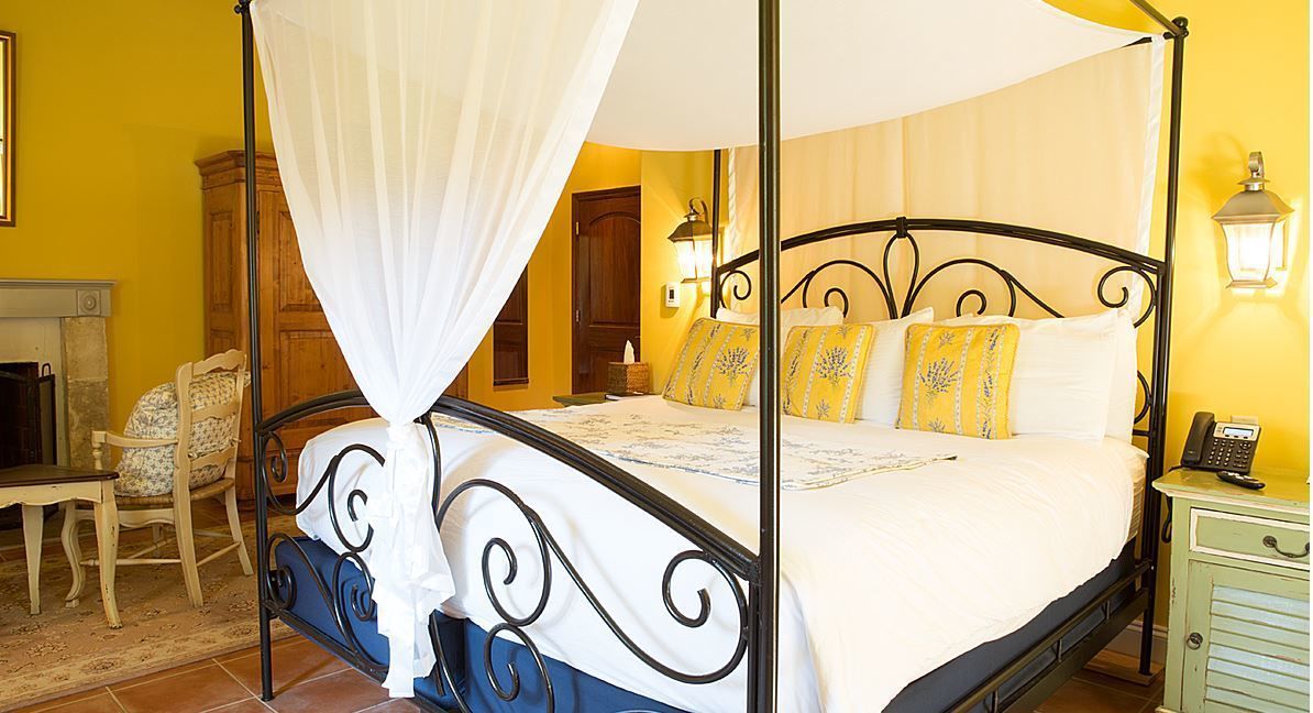 A four-poster bed with a canopy, a bedside table with a lamp, and a chair with a floral cushion.