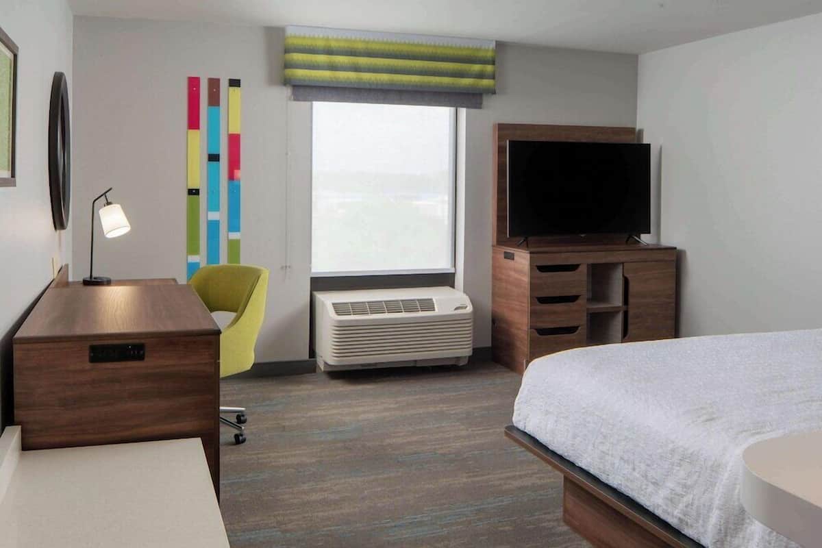 A hotel room with a bed, a desk, a chair, a TV, a wall-mounted air conditioner, and a window with a view.