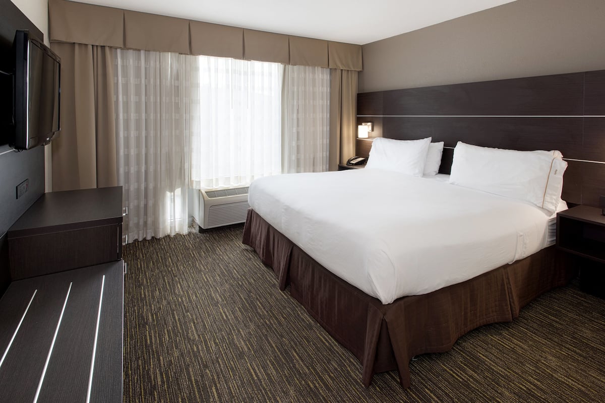 A hotel room with a large bed, a flat-screen TV mounted on the wall, a bedside table, and a window with curtains.
