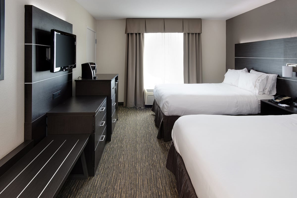 A hotel room with two beds, a TV mounted on the wall, a desk with a chair, and a window with curtains.