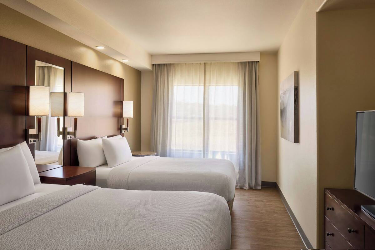 A hotel room with two beds, a large window with curtains, a flat-screen TV, and a wooden dresser.