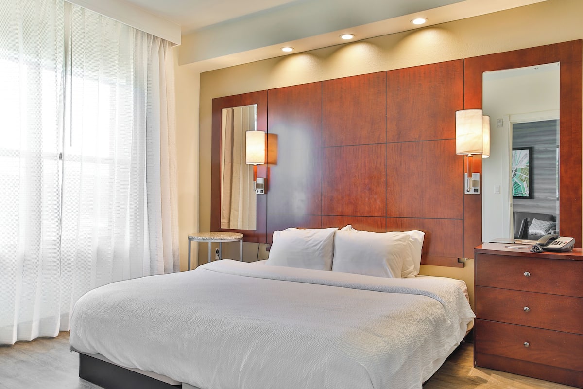 A hotel room with a large bed, a wooden headboard, a bedside table with a lamp, and a large mirror.