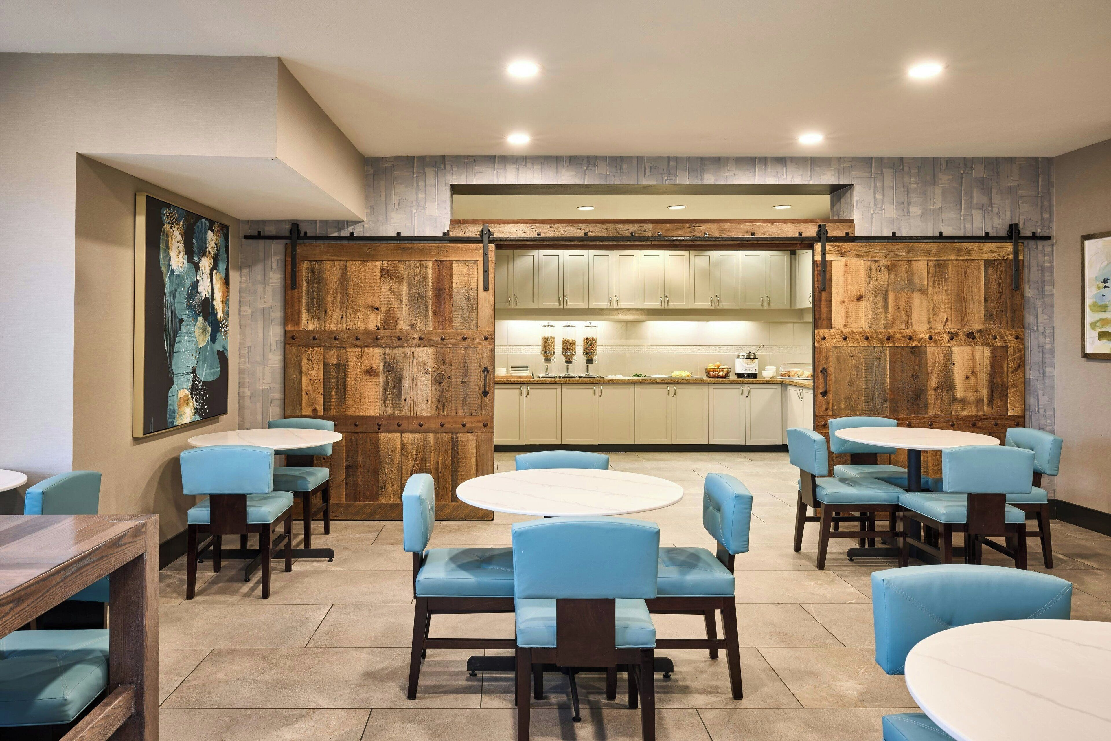 A dining area with round tables and blue upholstered chairs, a kitchen in the background, and wooden paneling on the walls.