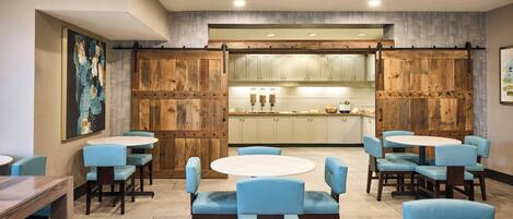 A dining area with round tables and blue upholstered chairs, a kitchen in the background, and wooden paneling on the walls.