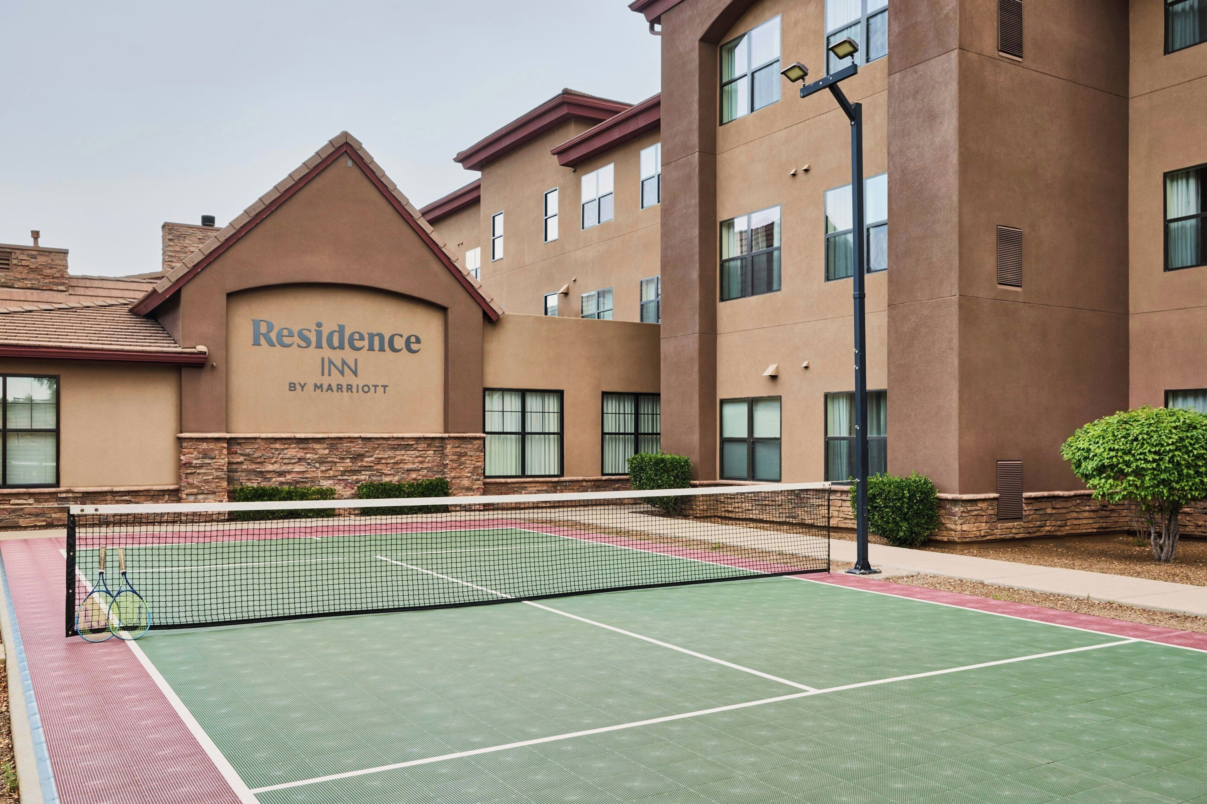 A tennis court with a net, a building with a sign reading 'Residence Inn by Marriott,' and a clear sky.