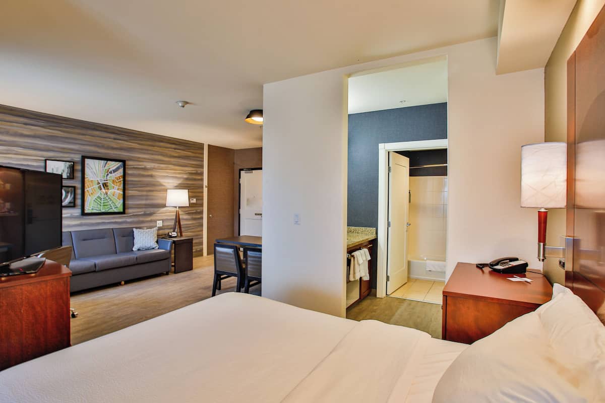 A hotel room with a large bed, a desk, a sofa, a TV, and a bathroom visible through an open door.