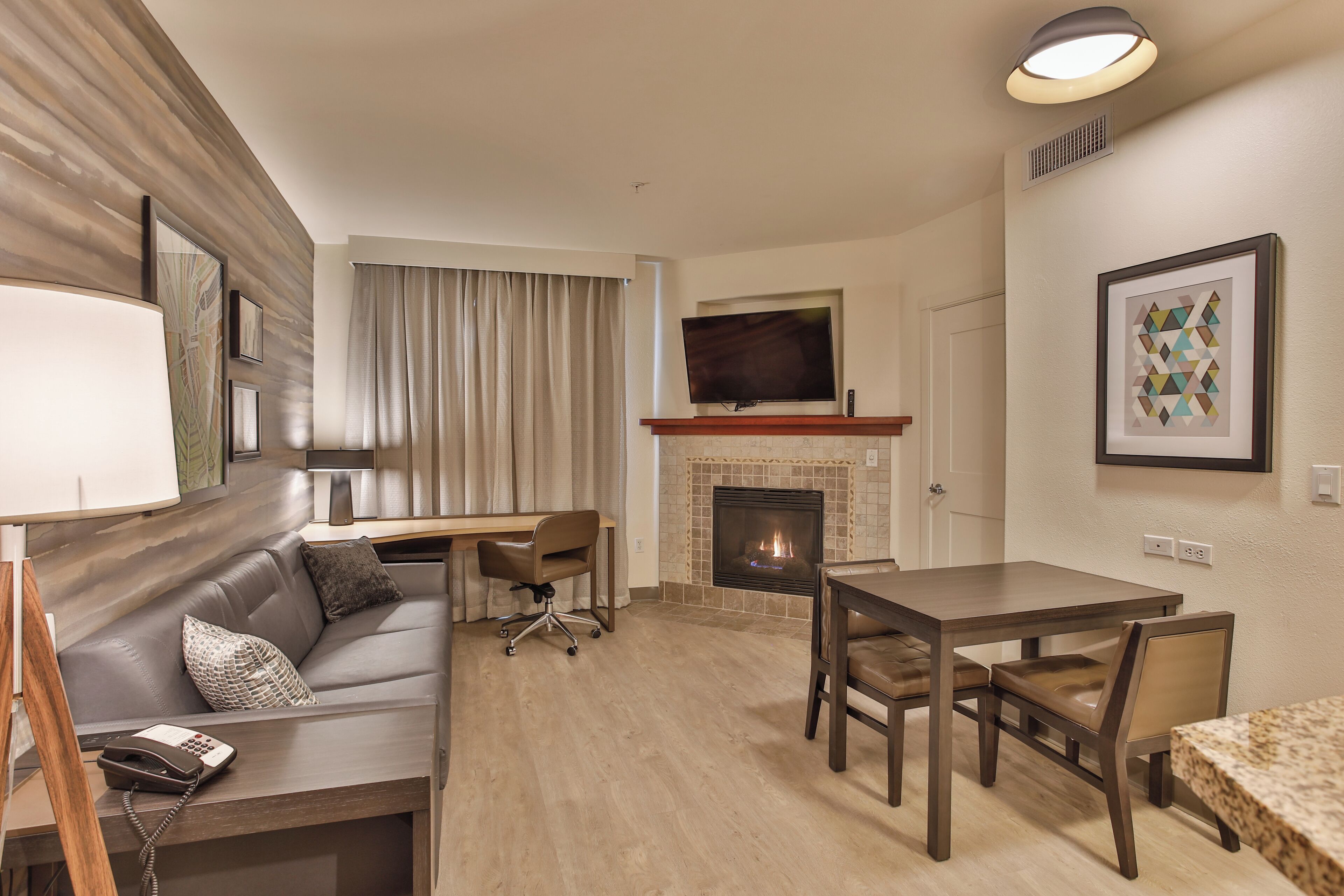Een moderne woonkamer met een open haard, een flatscreen televisie erboven, een eettafel met stoelen, een bank met een telefoon en een lamp op een bijzettafeltje, en een raam met gordijnen.