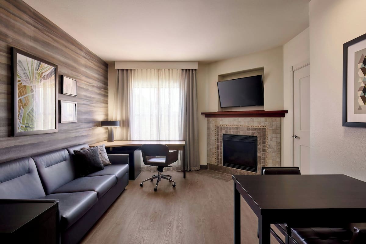 A modern hotel room with a leather sofa, a dining table, a fireplace, and a flat-screen TV mounted above it.