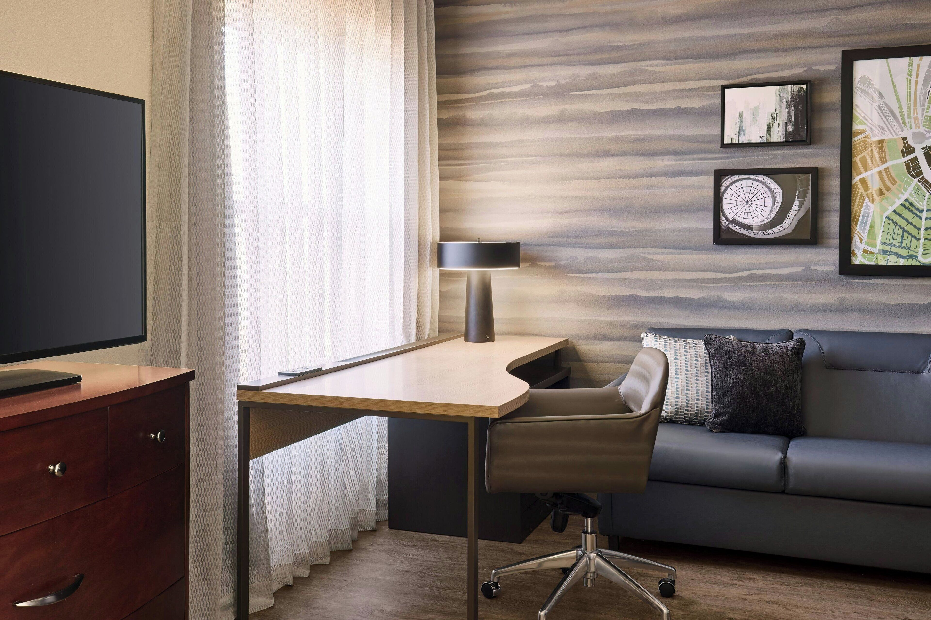 A modern hotel room with a wooden desk, a leather chair, a sofa, and a large flat-screen TV.