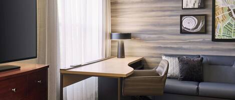 A modern hotel room with a wooden desk, a leather chair, a sofa, and a large flat-screen TV.