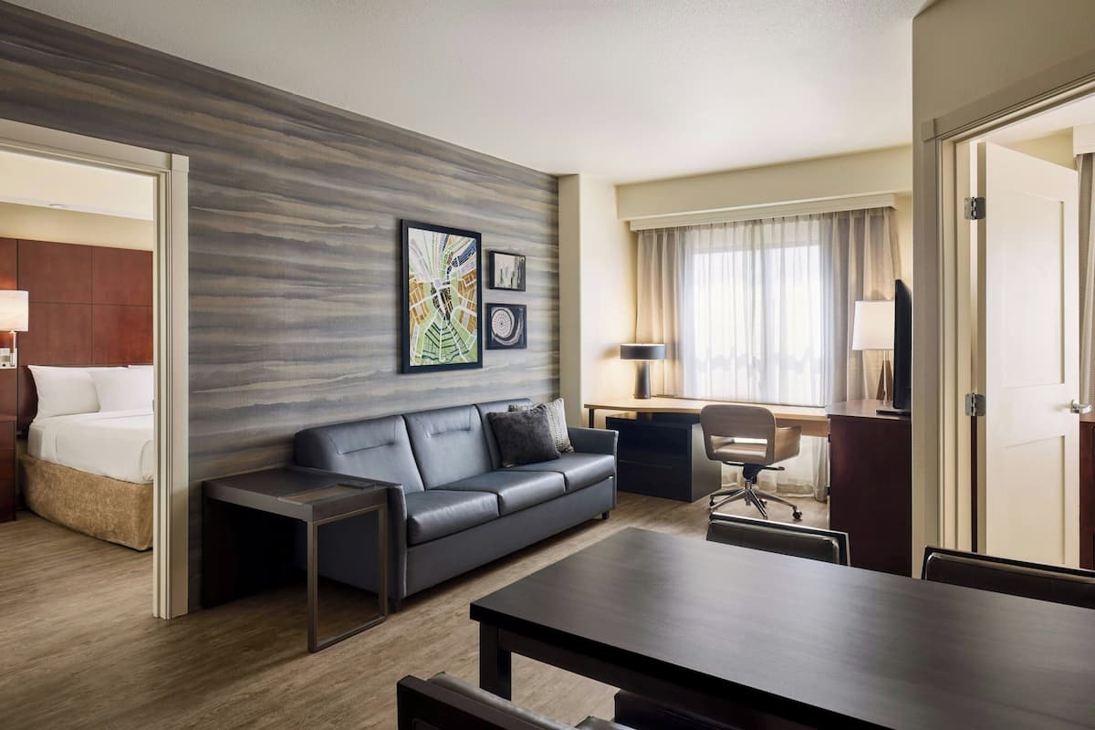 A modern hotel room with a grey leather sofa, a wooden coffee table, a desk with a chair, and a bed with a wooden headboard.