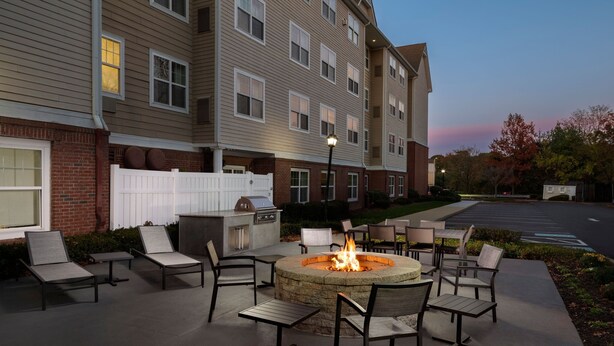 An outdoor patio with a stone fire pit surrounded by lounge chairs and tables, adjacent to a multi-story building with a brick facade and a white fence. - Residence Inn by Marriott Neptune at Gateway Center (Neptune City)