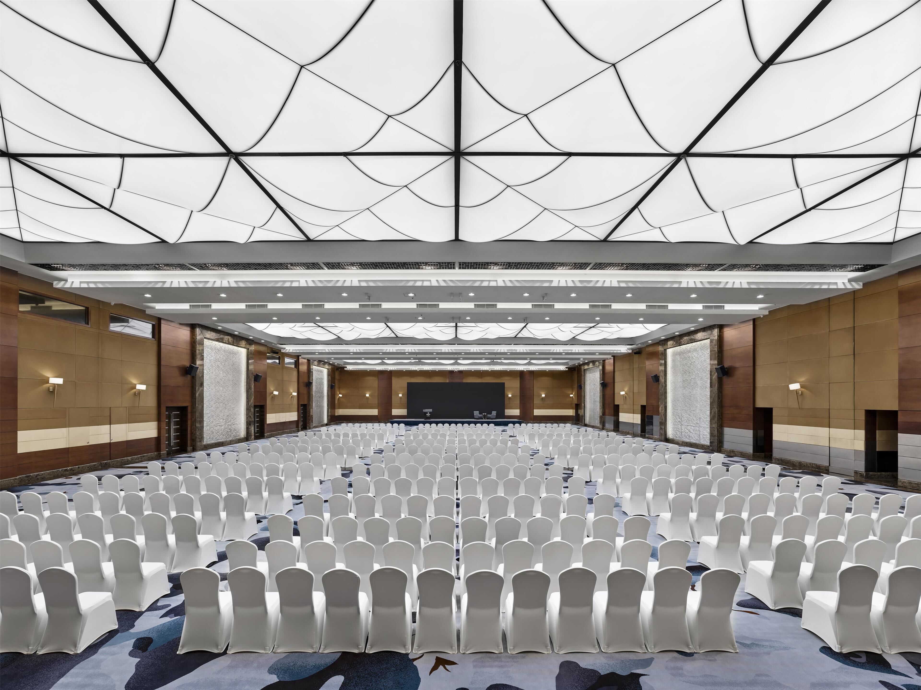 A large conference room with rows of white chairs, a stage, and a high ceiling with a geometric pattern.