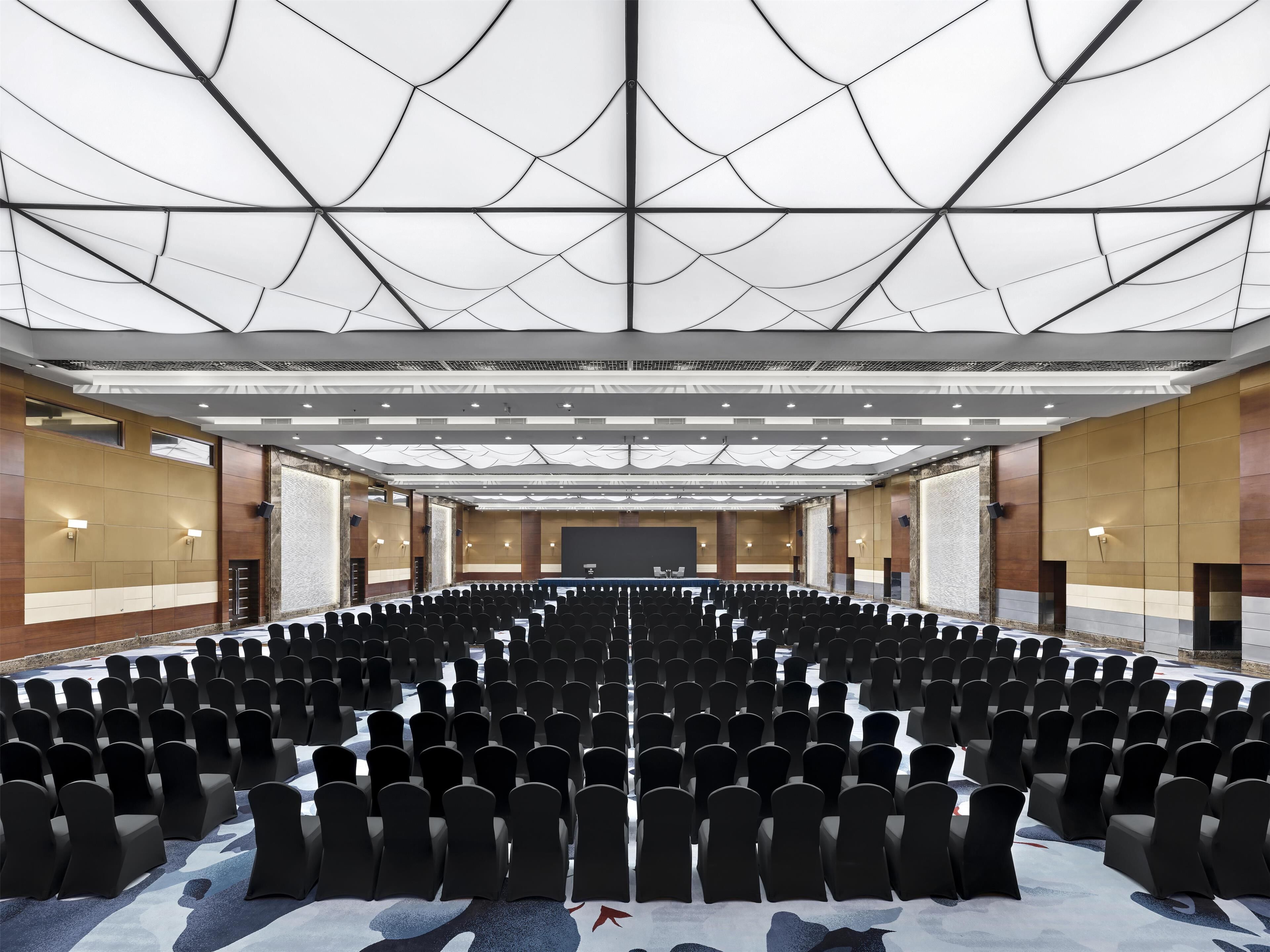 A large conference room with rows of chairs, a stage, and a high ceiling with a geometric pattern.