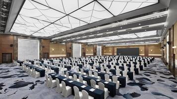 Une grande salle de conférence avec des rangées de tables et de chaises, un grand écran et un plafond haut orné d’un motif géométrique.