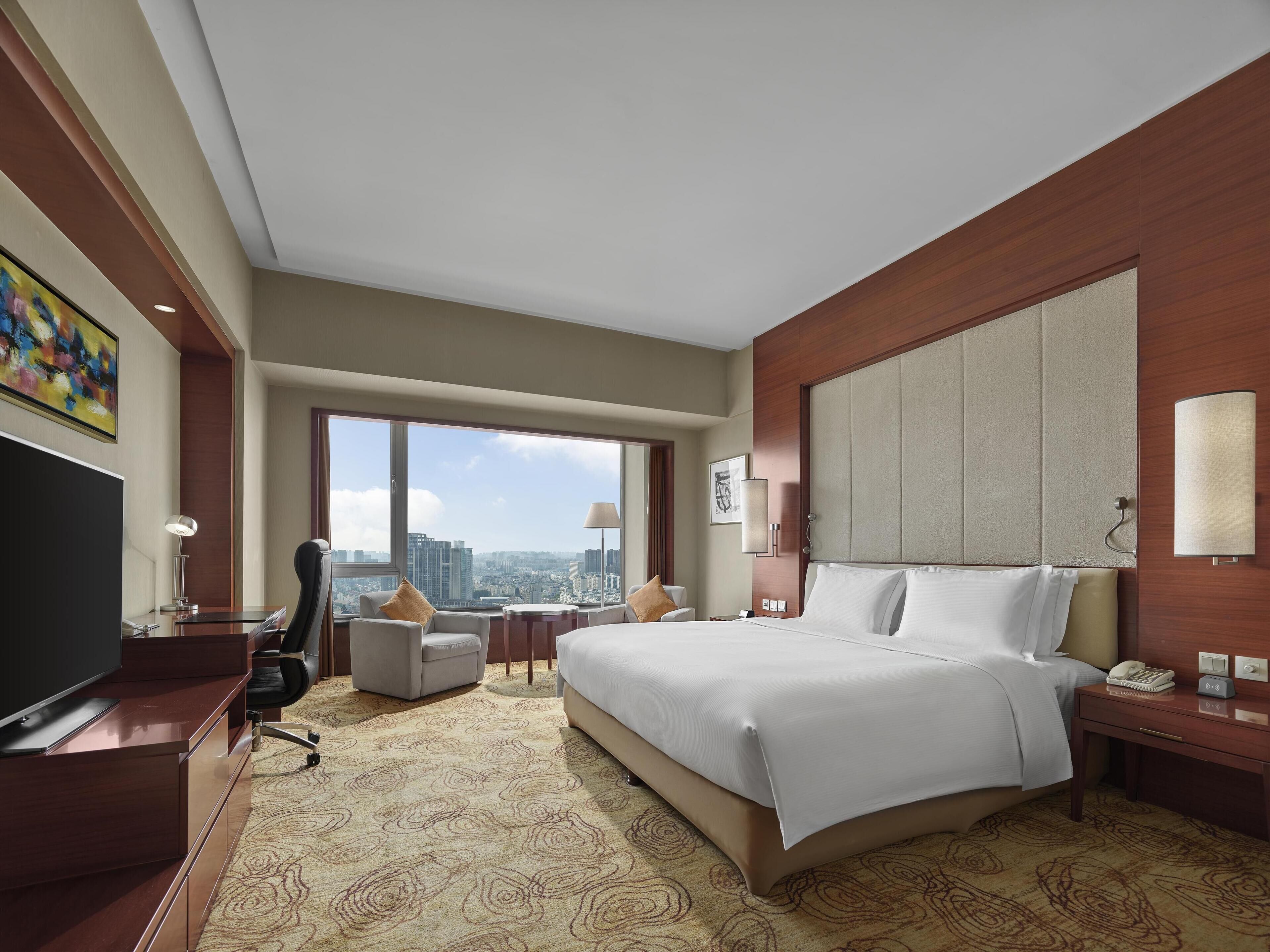 A hotel room with a large bed, a desk, a chair, a TV, and a city view.