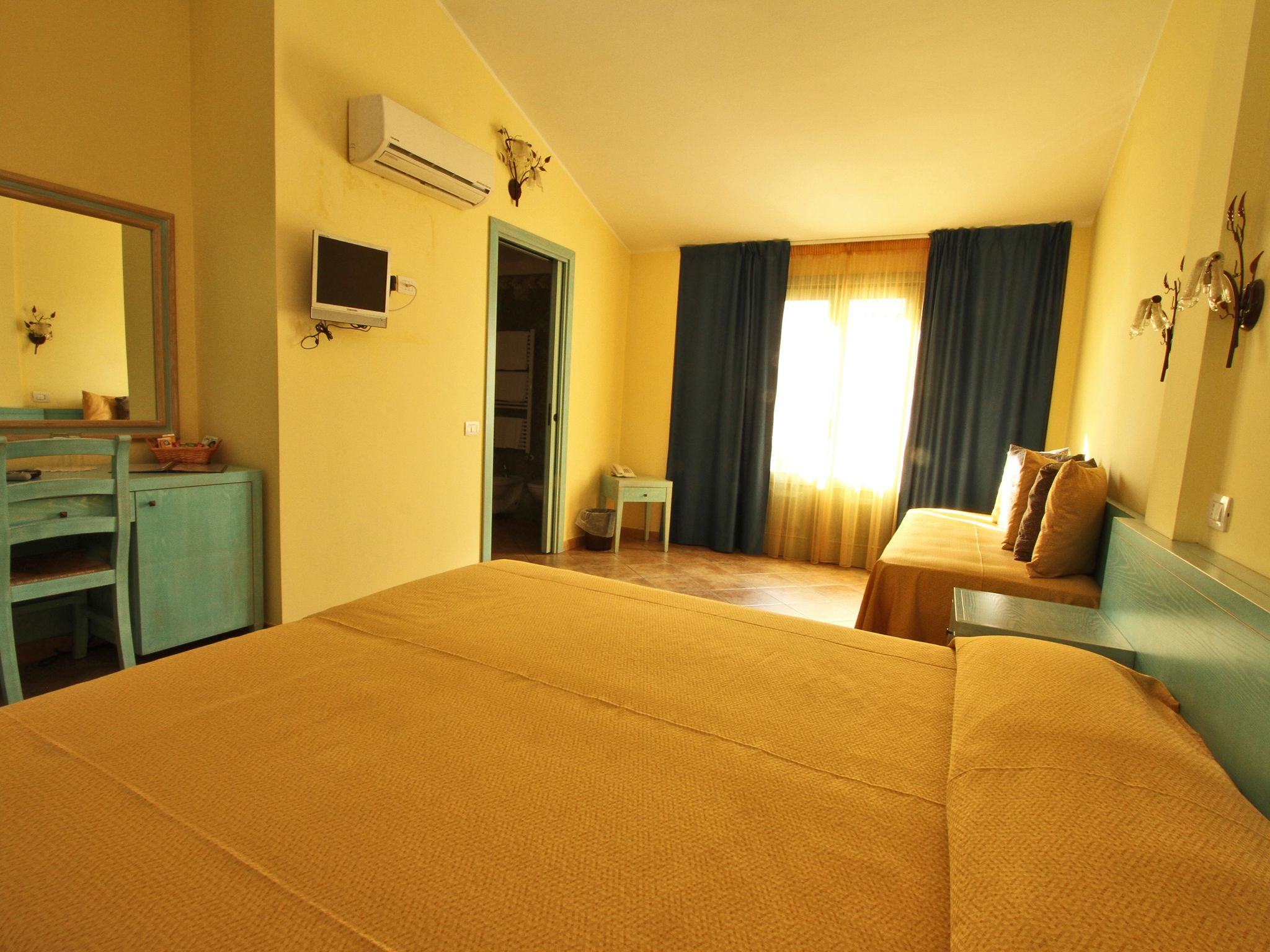 A hotel room with a large bed, a desk, and a chair. There is a window with curtains, an air conditioning unit, and a small table with a lamp.