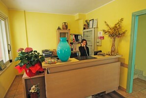 Un comptoir d’accueil avec un réceptionniste, un grand vase turquoise et une plante en pot ornée de fleurs roses.