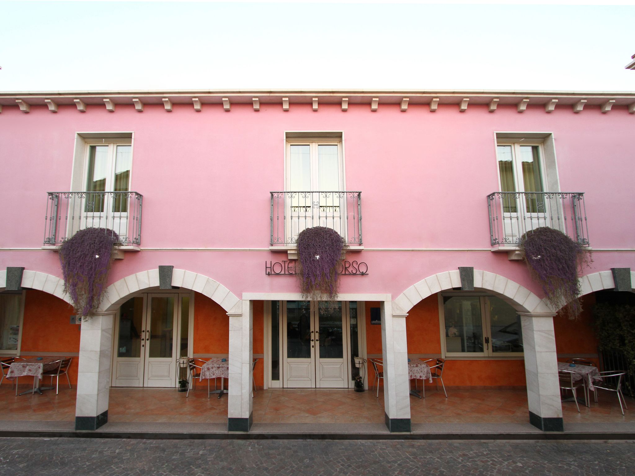 A pink building with a sign reading "HOTEL". There are three balconies with metal railings and hanging purple plants. The entrance has two doors with glass panels and a small table and chair outside.