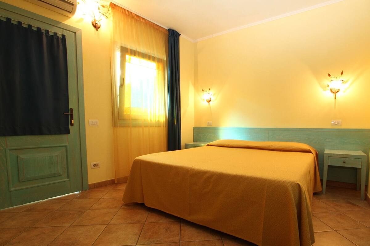 A hotel room with a bed, a nightstand, a window with curtains, and wall-mounted lights.