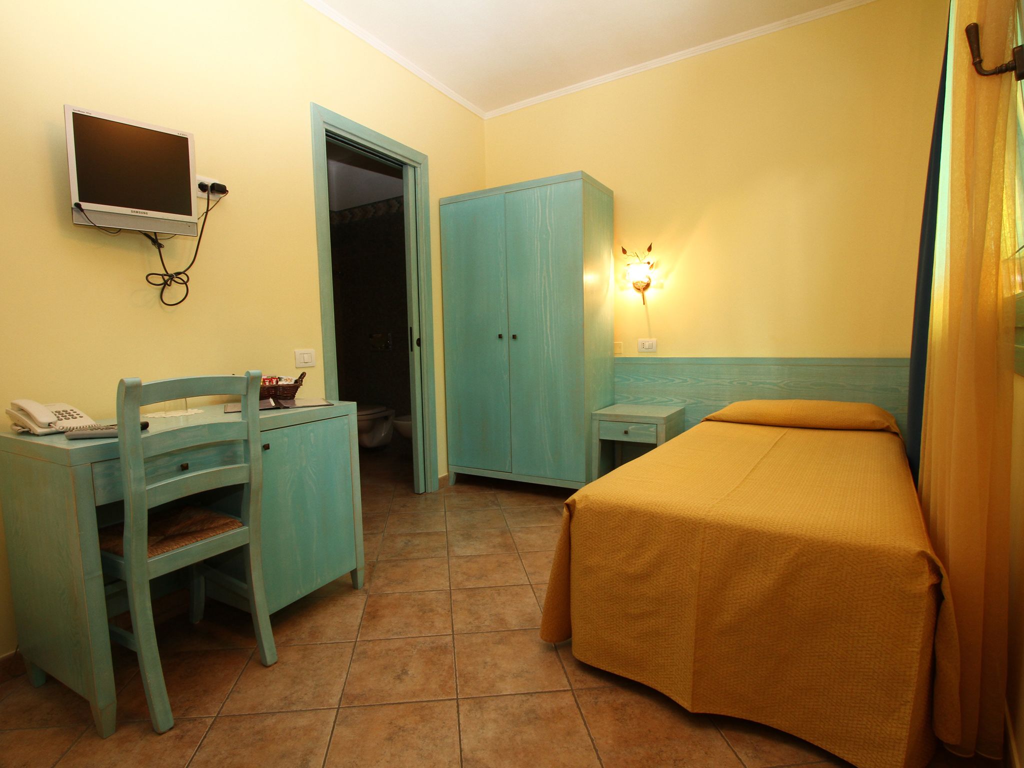 Ein Zimmer mit einem Bett, einem Schreibtisch mit Stuhl, einem Kleiderschrank, einem an der Wand befestigten Fernseher und einer Badezimmertür.