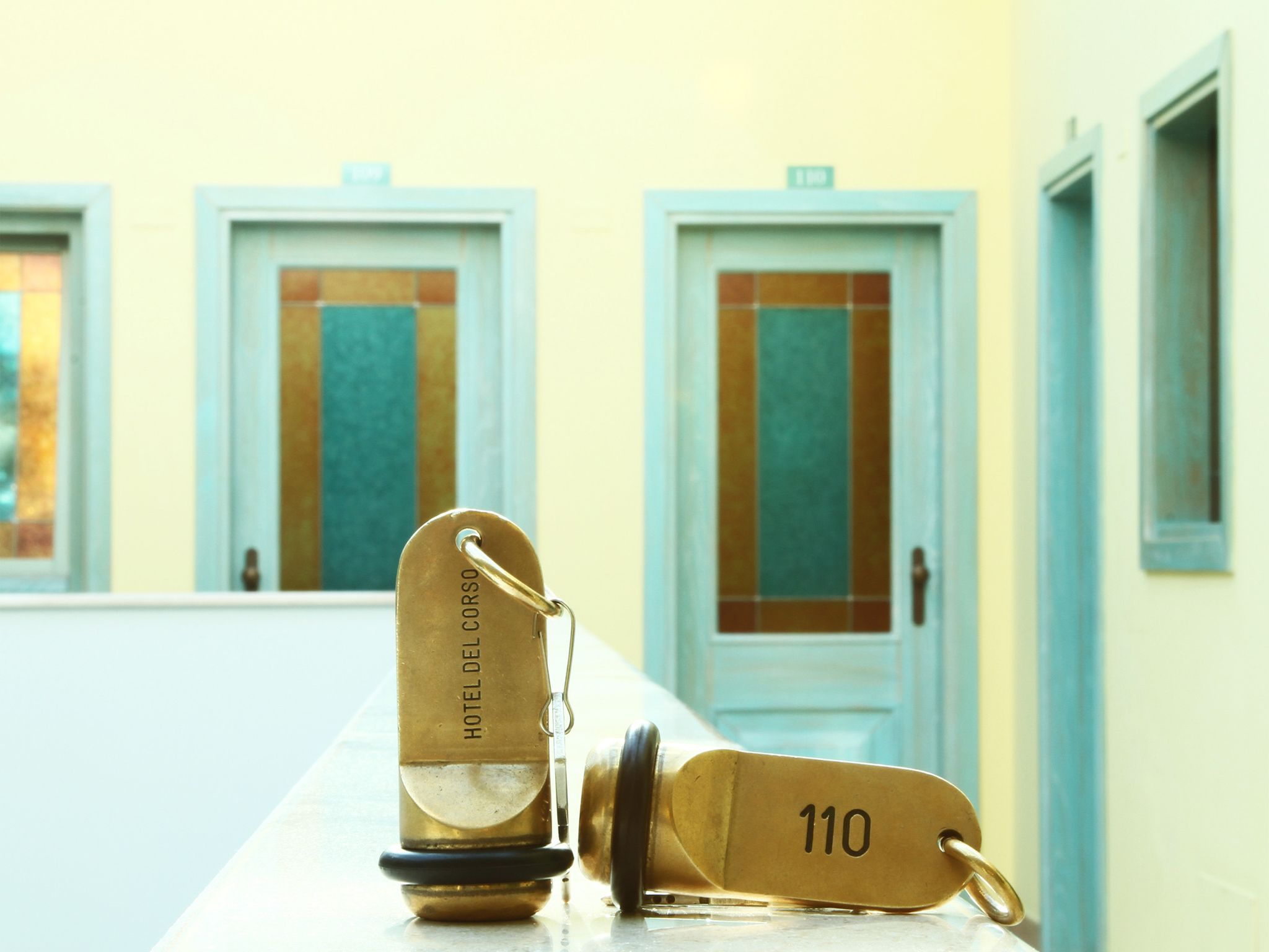 A brass key with "HOTEL DEL CORSO" and room number "110" engraved on it, lying on a counter with a row of doors in the background.