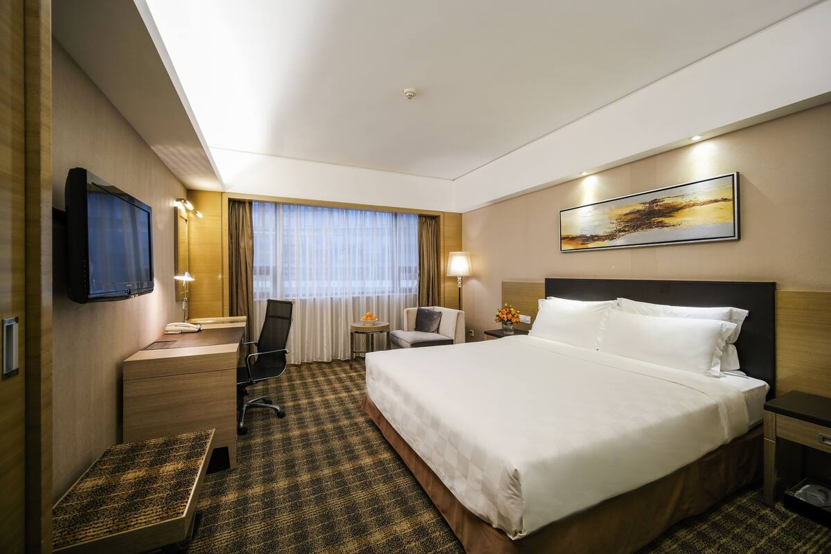 A hotel room with a large bed, a desk with a chair, a television, a window with curtains, and a painting on the wall.