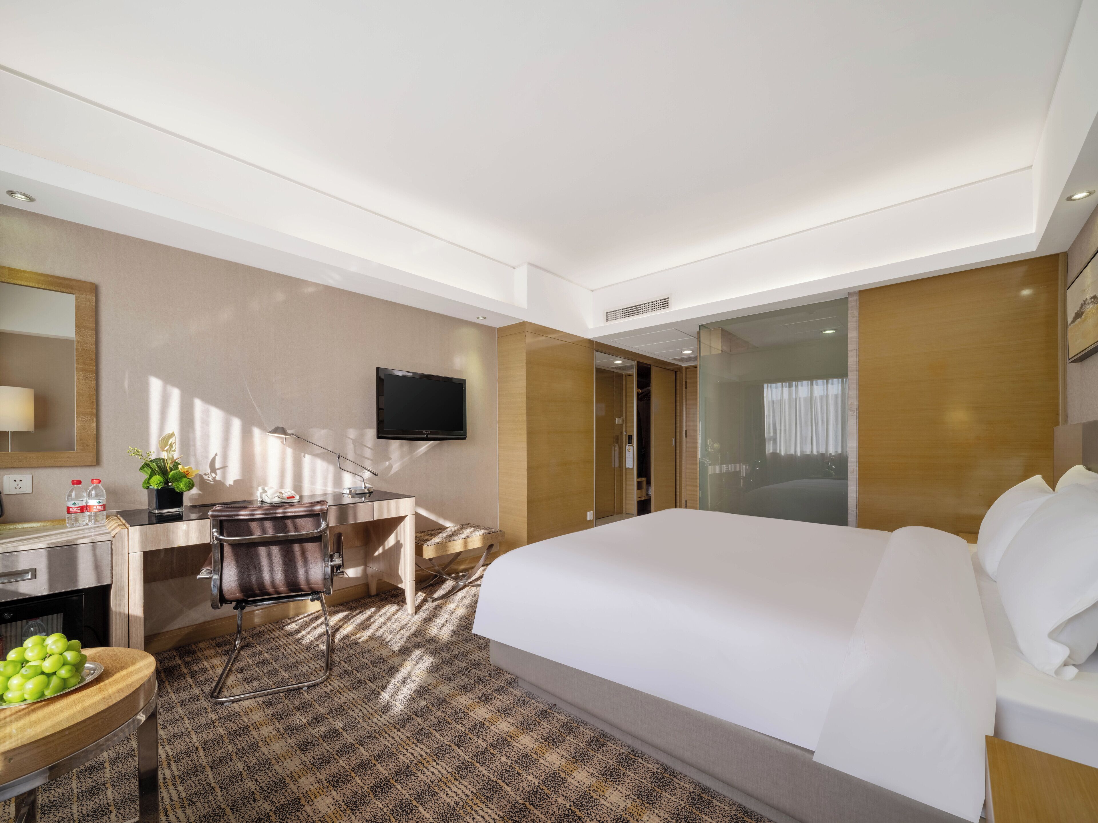 Een moderne hotelkamer met een groot bed, een bureau met stoel, een flatscreen televisie aan de muur en een klein tafeltje met een schaal fruit.
