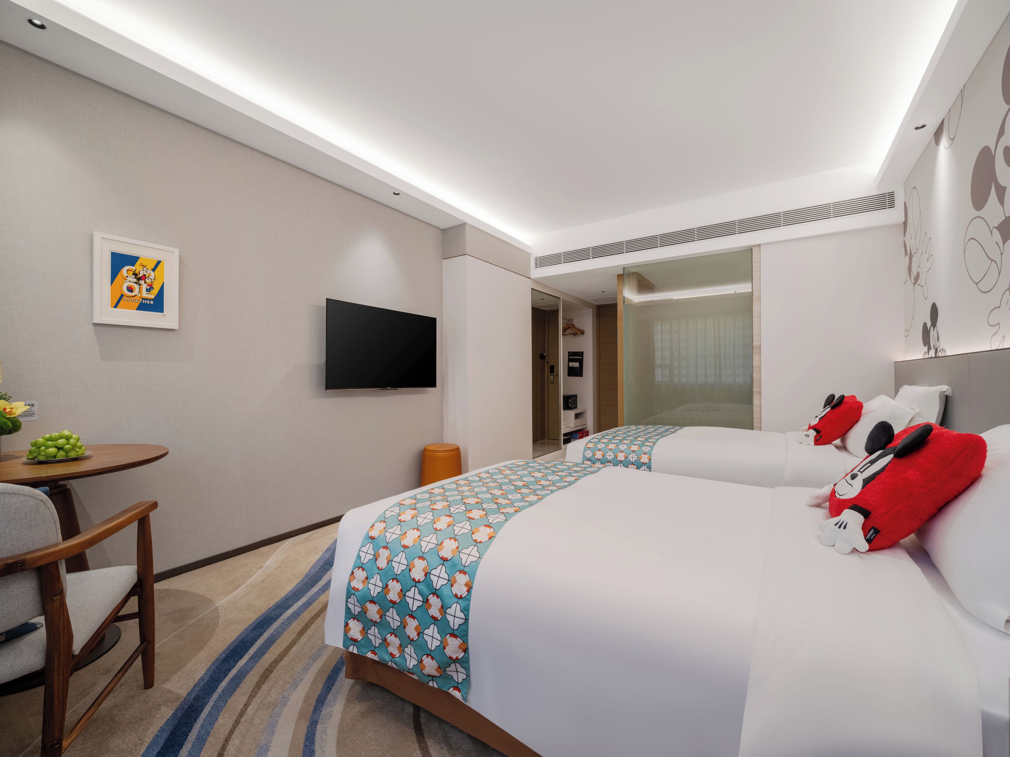 A hotel room with two beds, a TV, a desk with a chair, and a small table with a fruit bowl.
