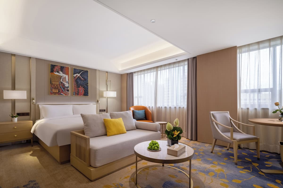 A modern hotel room with a large bed, a sofa, a dining table, and a chair. There are two paintings on the wall, a lamp on the nightstand, and a flower vase on the table.