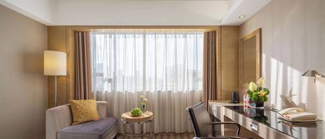 A hotel room with a sofa, a desk, a chair, a small table with a vase of flowers, and a large window with curtains.