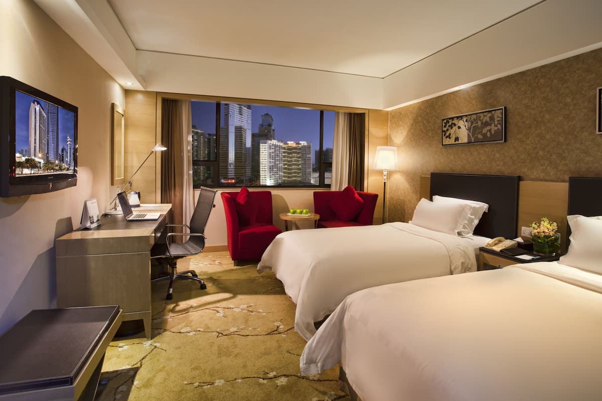 A hotel room with two beds, a desk, a chair, a TV, and a view of the cityscape.