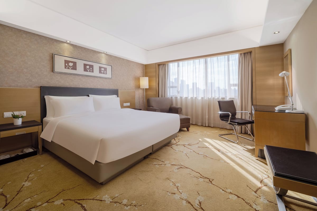 A hotel room with a large bed, a desk, a chair, a lamp, and a view of the city through the window.