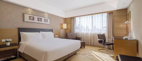 A hotel room with a large bed, a desk, a chair, a lamp, and a view of the city through the window.