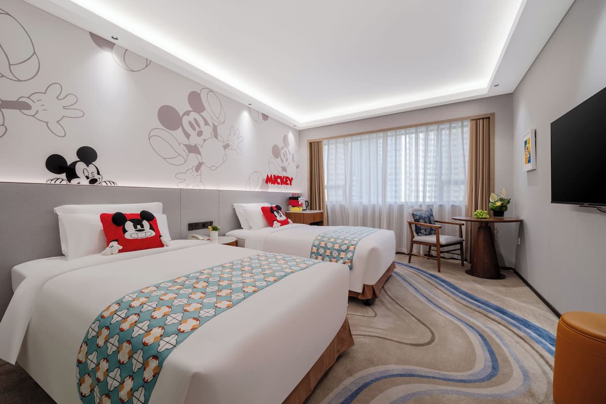 A hotel room with two beds, a desk, a TV, and Mickey Mouse-themed decor.